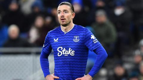 Everton's Jack Grealish stood on a fotball pitch wearing a blue Everton FC shirt with his hands on his hips