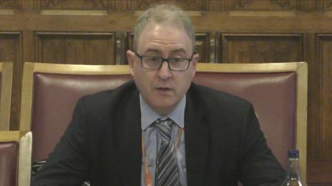 Geoff Darch with thinning hair and wearing glasses and a black suit with blue shirt and grey tie sits in a large chair while giving evidence to the select committee. 