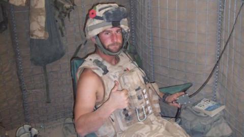 Major John Whiteman in uniform in a military shelter with a helmet on and sitting on a green chair.