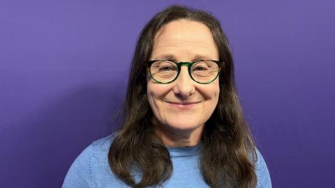 A woman stood in front of a purple wall. She has long brown hair and glasses and is wearing a pale blue top