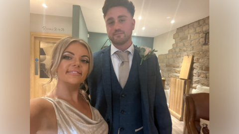 A young woman with blonde hair and a glittery dress, stands next to a young man in a navy suit inside a pub