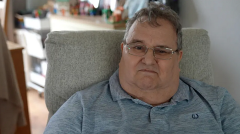 A close-up of David Chapman, wearing a grey polo shirt and glasses, as he sits in an armchair