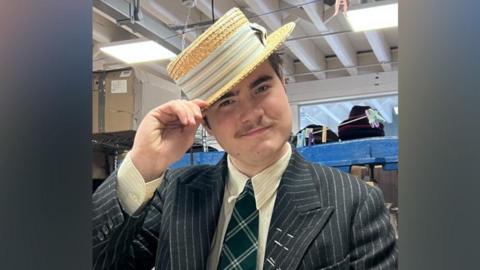A person in a dark pinstripe suit, cream shirt, and green plaid tie stands indoors holding a straw boater hat. Behind them are shelves filled with hats and boxes in a workshop‑like space with exposed beams and fluorescent lighting.