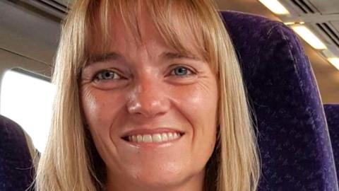 Sandie Peggie, a woman with shoulder-length blonde hair, smiles at the camera. She appears to be sitting on public transport.