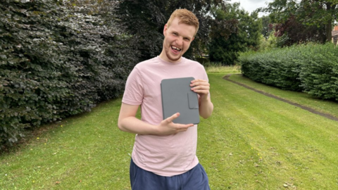 Man wearing a pink T-shirt stands in a back garden, which has hedges either side and a lawn, while holding an iPad with a grey cover
