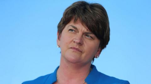 Arlene Foster stares upwards to her right. She has short dark hair and is wearing a blue suit jacket