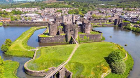 A general view of Caerphilly castle from the air, with the building situated in the centre of the picture surrounded by a moat and a bridge crossing it, and the town in the background.