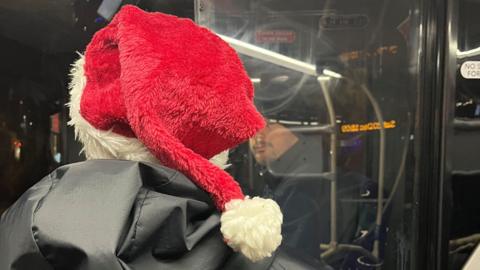 A police officer wearing a santa hat, speaking to a bus driver who is sitting behind glass.