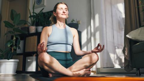 A woman in a grey top meditating on the floor of a room with lots of plants and a cupboard. She has her legs crossed, her eyes closed and her hands on her knees.