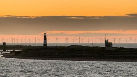 A view of a thin spit of land sandwiched between the North Sea and the Humber Estuary featuring a lighthouse. Multiple wind turbines can be seen in the distance. The sky is orange and cloudy.