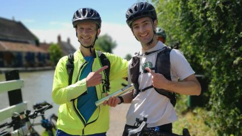 Damian Hall and David Starley. Damian is wearing a bike helmet and a yellow neon jacket. He has his arm around David and is pointing at him, smiling. David is also wearing a helmet and a white t-shirt, smiling at the camera.