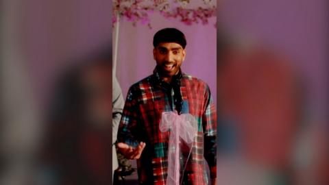 A man wearing a red tartan shirt, mid speech, in front of mic with a pink bow tied to it.