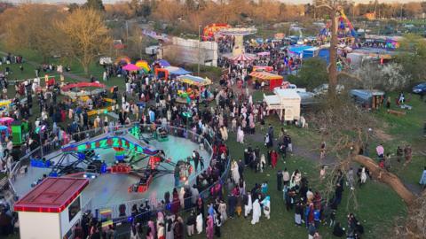 A large group of people at an Eid event in Luton, taken from high up, showing a fairground, stalls, people and trees in a park. 