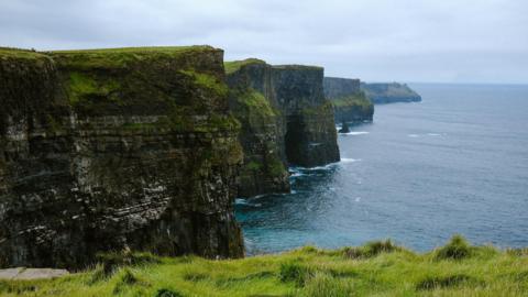 A view of the Cliffs of Moher on a cloudy day with a calm sea below.  The tall, rugged cliffs jut out at angles to the sea. 