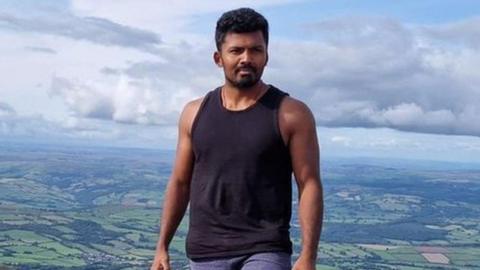Mohananeethan Muruganantharajah is wearing a black vest-top and jeans while standing high on hillside overlooking a panoramic countryside view