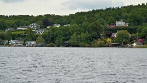 Eastern shore of Windermere in the Lake District. It has a few houses and boat houses dotted in between a forest. 