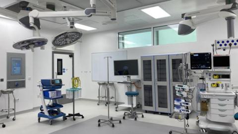 The inside of one of the surgical theatres. It is a white, starkly lit room with lots of medical equipment on wheels and overhead lighting. 