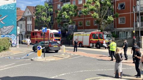 Two fire engines parked inside a cordon, with blue and white police tape securing a large section of the main road. 