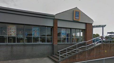 The Aldi store on Risedale Road. The supermarket is made of grey and red bricks. A set of stairs leads to the entrance where plants and flowers are displayed on racks.