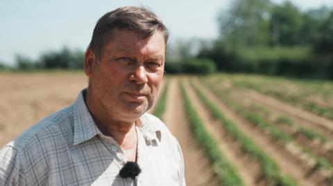 Chris Suckling standing outside in a field. There are rows of crops growing in the background behind him, which is blurred. He is wearing a checked shirt and is looking directly at the camera.