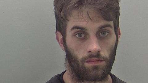 A man with dark hair and a beard is looking at the camera.