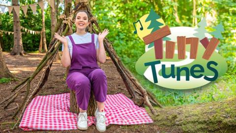 Lisa sat singing in a woodland play area, smiling and signing, she is wearing purple dungarees. The logo 'Tiny Tunes'.