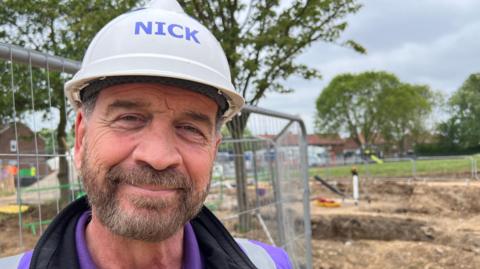 TV presenter Nick Knowles smiles into the camera. He has a beard and is wearing a white hard hat, purple t-shirt, black top and a purple high-vis vest. He is standing in front of a building site and a metal fence. Trees and houses are visible in the distance.