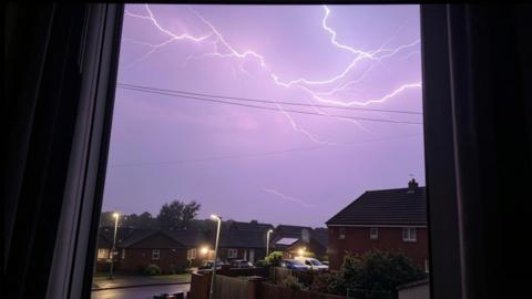 Lightning in the purple sky over a housing estate - through a window frame with curtains