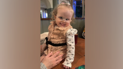 Ivy, a small girl, sits on a table smiling at the camera. She has glasses and curly, light blonde hair tied back in a white bow. She is wearing a peach coloured, furry gilet with a black ribbon tied around it, and a white, long-sleeved top with black ribbons on. 