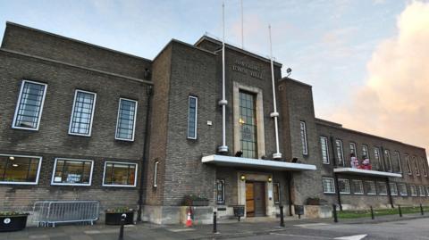 A landscape shot of Havering Town Hall. It is a wide, dark brown brick building with long windows, in an international modern architectural style. 