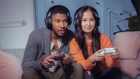 Stock image shows a young couple on a sofa, wearing over-the-ear headphones and holding Xbox controllers. The young man on the left looks intense and leans forward, concentrating. The female player on the right looks more relaxed.
