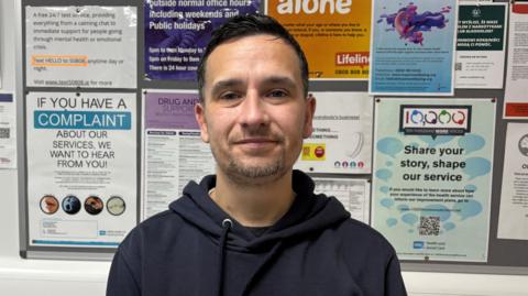 Michael Main is wearing a navy hoodie. He has short black hair and grey facial hair. .He is standing in front of a notice board with lots of coloured posters on it.