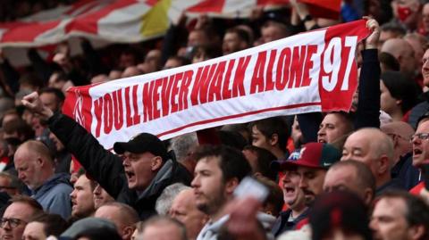 A crowd of Liverpool Football Club supporters holding a banner which reads 'You'll Never Walk Alone 97' 