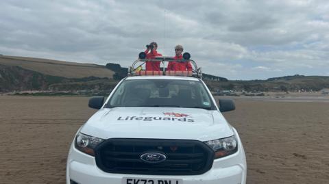 Two young male lifeguards in red jackets stand on the back of a white RNLI branded truck. They are on a sandy flat beach, with hills behind.