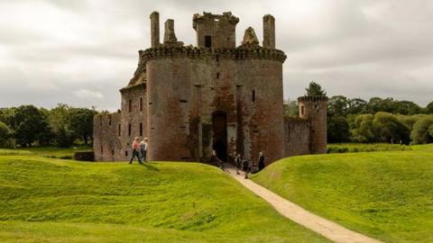 The remains of a 13th Century Castle - Caerlaverock - stand in a hilly green landscape. A few visitors walk around the outside and across a bridge leading over a moat which sits around the ruined structure.