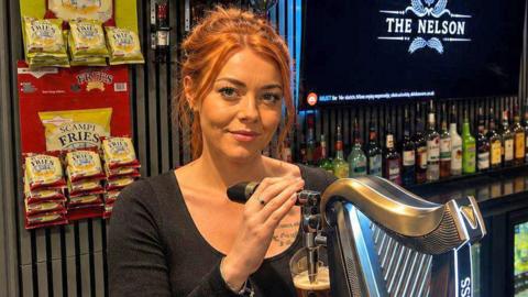 A woman with ginger hair stood behind a bar with various brewery logos displayed on taps. There is a screen in the background with the pub name - The Nelson - displayed on it.