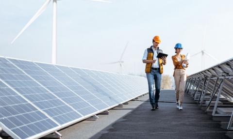 Two workers wearing hard hats inspect rows of solar panels, with three wind turbines in the background.