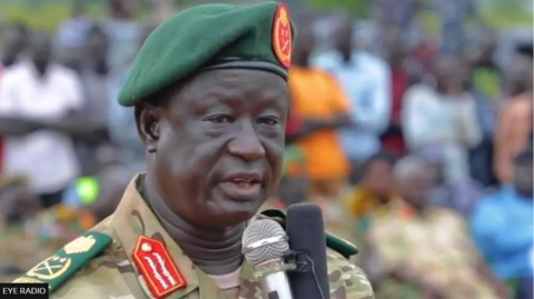 General Akol Koor Kuc wears army regalia, including a green beret, as he speaks into a microphone. A crowd can be seen behind him