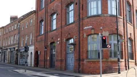The former Gateshead Railway Club on Wellington Street. It is a large three-storey building made out of dark orange brick with blue-grey window frames. It sits on a corner by some traffic lights, with a curved wall as it bends between the two roads.