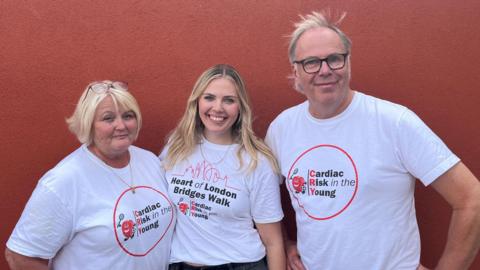 Two women and a man are stood together in front of a red wall. They are all smiling and wearing white T-shirts that display a Cardiac Risk in the Young logo. The first woman, on the left of the picture, has short blonde hair and a pair of glasses on top of her head, the second woman, in the centre of the picture has long blonde hair and a T-shirt that also displays the words: Heart of London Bridges Walk. The man on the right of the picture has short grey hair and is wearing glasses. 