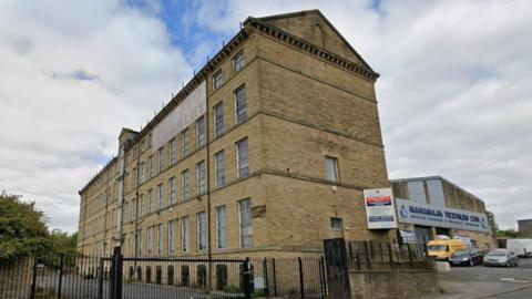 An exterior photo of a four storey mill building. It sits behind black metal railings and gates. It looks neat and well kept. A huge banner hanging from the top of the building says 'To Let'. To the right is a business premises, Maharaja Taxtiles. There are several cars and vans parked in front of the textile shop.