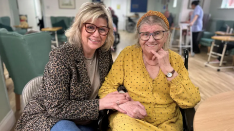 Leonie Williams and her mum sit together in the living room of a care home with their arms linked, smiling towards the camera. Leonie is wearing jeans, a top and a leopard print jacket, while her mum wears a yellow spotted dress