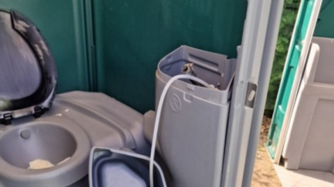 A damaged temporary toilet. The toilet is grey and part of it has been vandalised. The toilet lid is open. The back of the toilet block is green.