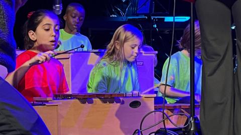 Two young girls, one dressed in red and the other in green, are sitting on the floor of a stage and are playing on xylophones