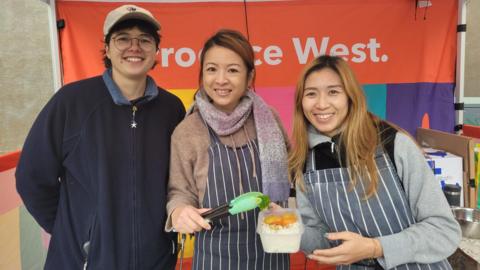 Mel Fu, Jenny Luk and Vicky Yung (L-R) are standing next to each other at a food stall. They are all smiling, with the two women on the right wearing striped aprons.