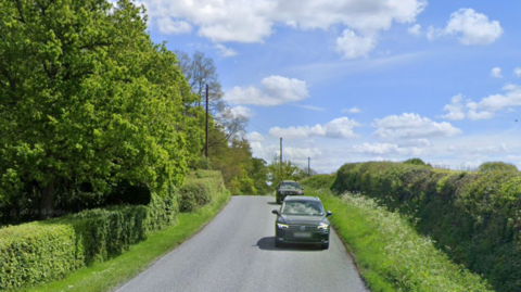 A google street view imagine of a country lane on a sunny day, with two cars travelling towards the viewer 