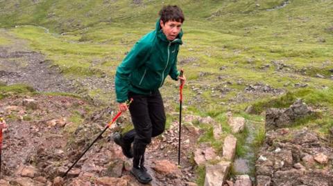 Trish Patterson, a woman with short hair walking with two walking sticks on a rocky terrain. She is wearing black trousers and a green coat and appears out of breath as she looks to the camera.
