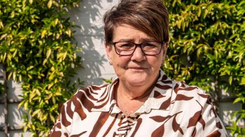 A head and shoulders picture of Tracy Page, wearing a brown and cream patterned top, sitting in front of a wall with climbing plants.
