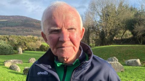  Kevin Murphy wearing a navy O'Neills branded coast and green zip jumper underneath. he is standing in front of rocks and grassy hills 