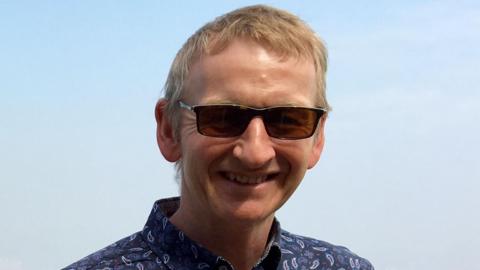 Neal Doggett has short blonde hair and is smiling brightly at teh camera. he is wearing black sunglasses and a blue shirt.